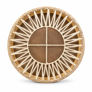 May include: A circular frame drum featuring a light brown skin head stretched across a wooden hoop. The back of the drum is secured with a complex lacing pattern made of thin, cream-colored cord that forms a star-like design around a central cross.