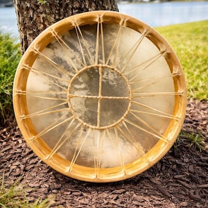 May include: A round, wooden drum with a natural hide drumhead. The drum is held together with woven cord and has a cross design in the center. The drum is resting against a tree trunk, with a lake in the background.
