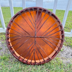 May include: A round, handcrafted drum with a natural leather drumhead and a dark brown wooden frame. The drumhead is a warm orange color, and the frame is intricately laced with dark brown leather. The drum is resting on green grass.