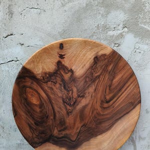 Pode incluir: Tábua de servir oval de madeira com um rico padrão de grão em espiral em tons de castanho e bege. A tábua tem uma forma natural e orgânica e é provavelmente feita de madeira de lei. Repousa sobre uma superfície de madeira envelhecida.