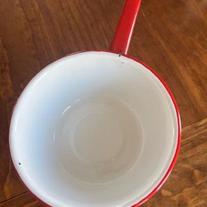 May include: A small, white enamel saucepan with a red rim and handle. The handle has a hole for hanging. The interior of the pan is white, and the exterior is red. The saucepan is set against a wooden surface.