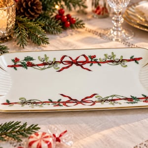 Peut inclure: Un plateau de service rectangulaire en porcelaine blanche avec des bords festonnés et une bordure dorée. Le centre présente un motif répétitif de rubans rouges, de feuilles de houx vertes et de baies blanches, symétrique sur les côtés.