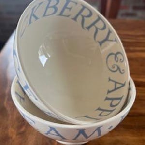 May include: Two off-white ceramic bowls stacked on a wooden table. The bowls have a light blue rim and the words "Blueberry & Apple Jam" printed in light blue. The bowls are angled, showing the interior.