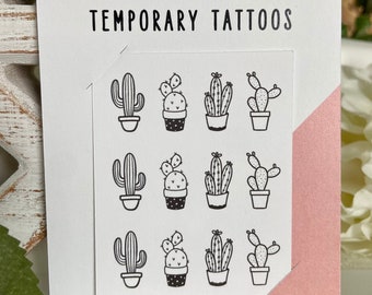May include: A white card with the text "TINY TATTOO COMPANY TEMPORARY TATTOOS" and a series of black cactus designs. The card has a pink accent and is surrounded by greenery.