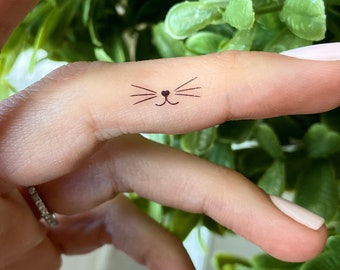 May include: A small, black cat face tattoo on a finger. The tattoo features a simple design of whiskers, a nose, and a heart-shaped mouth. The finger is held up against a background of green foliage.