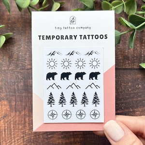 May include: A white rectangular card labeled "tiny tattoo company" and "TEMPORARY TATTOOS" featuring a grid of black ink designs including mountain ranges, suns, bears, pine trees, and compasses printed on a white backing sheet.