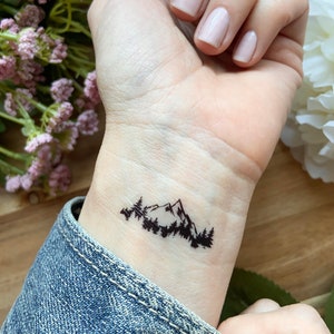 May include: A black mountain and forest temporary tattoo on a wrist. The tattoo features a silhouette of a mountain range with trees in the foreground. The wrist is against a denim jacket.