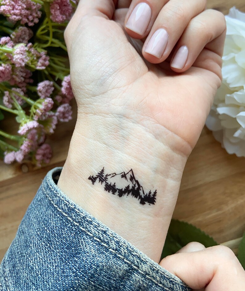 May include: A black mountain and forest temporary tattoo on a wrist. The tattoo features a silhouette of a mountain range with trees in the foreground. The wrist is against a denim jacket.