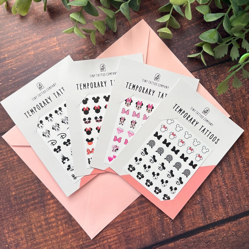 May include: Four white rectangular cards labeled Tiny Tattoo Company Temporary Tattoos, each featuring a grid of small black, pink, and red character-themed icons including mouse ears, bows, hearts, and diamonds, arranged on a light pink envelope.