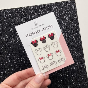 May include: A sheet of temporary tattoos on a white backing card labelled Tiny Tattoo Company Temporary Tattoos. The sheet features twelve small black and white mouse head icons, some with solid black ears and red bows, others as black outlines with red bows.