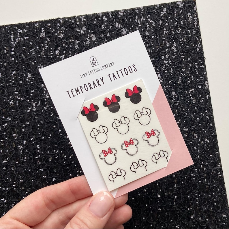 May include: A sheet of temporary tattoos on a white backing card labelled Tiny Tattoo Company Temporary Tattoos. The sheet features twelve small black and white mouse head icons, some with solid black ears and red bows, others as black outlines with red bows.