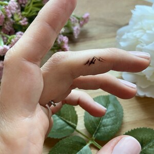 May include: A black mountain range temporary tattoo on a finger. The tattoo features three peaks with detailed shading, creating a minimalist design. The skin tone is fair, and the background includes soft pink and white flowers.