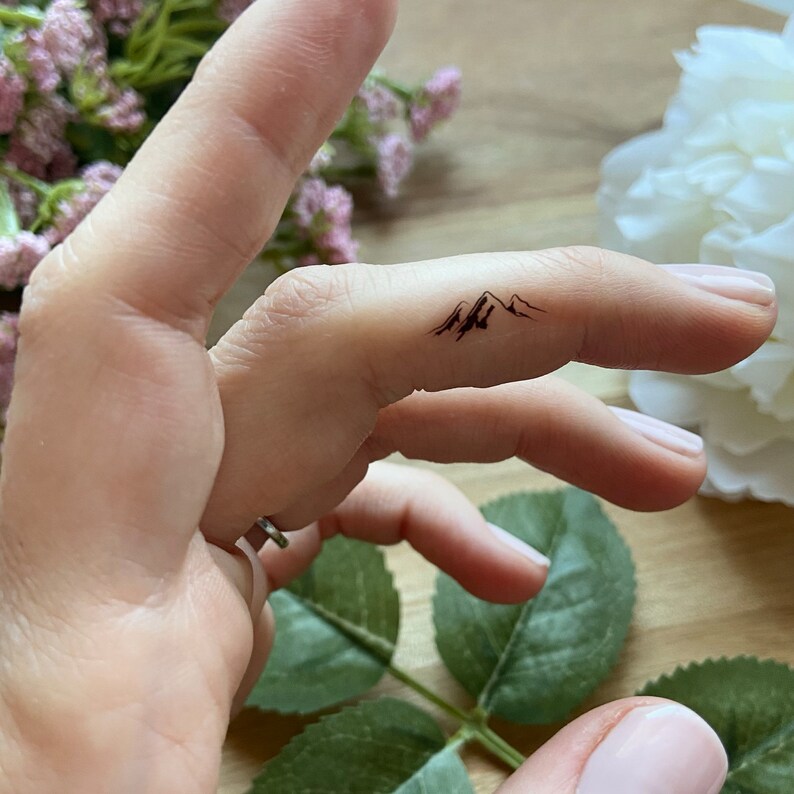 May include: A black mountain range temporary tattoo on a finger. The tattoo features three peaks with detailed shading, creating a minimalist design. The skin tone is fair, and the background includes soft pink and white flowers.