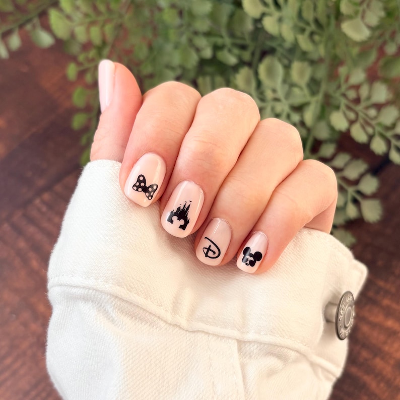 May include: Fingernails painted with a light pink polish featuring black nail art decals. The designs include a polka dot bow, a castle silhouette, a stylized letter D, and a Mickey Mouse head icon. The nails are clean and well-manicured.