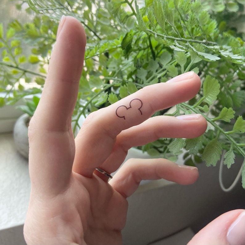 May include: A small, minimalist black ink tattoo on the side of a finger featuring the simple outline of a circular head shape with two rounded ears, creating a recognisable silhouette of a classic cartoon character icon.