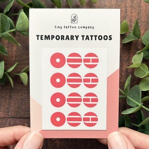 May include: A white rectangular card labeled tiny tattoo company TEMPORARY TATTOOS featuring a sheet of twelve small red circular designs arranged in four rows of three, including solid circles with center dots, horizontal stripes, and split bars.
