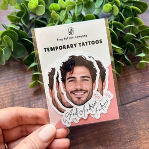 Puede incluir: Un paquete de pl&aacute;stico transparente que contiene tatuajes temporales con el retrato de un hombre de cabello oscuro y barba. El texto dice tiny tattoo company TEMPORARY TATTOOS y el nombre And Andrew con peque&ntilde;os iconos de corazones rosas.