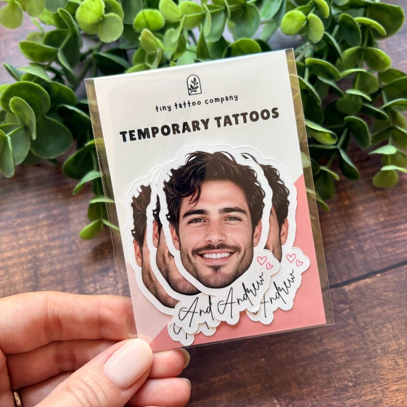 Puede incluir: Un paquete de pl&aacute;stico transparente que contiene tatuajes temporales con el retrato de un hombre de cabello oscuro y barba. El texto dice tiny tattoo company TEMPORARY TATTOOS y el nombre And Andrew con peque&ntilde;os iconos de corazones rosas.