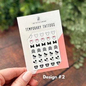 May include: A sheet of temporary tattoos on a white backing card labeled Tiny Tattoo Company Temporary Tattoos. The sheet features rows of small black and white icons including Mickey Mouse ears, bows, geometric spheres, and character faces.