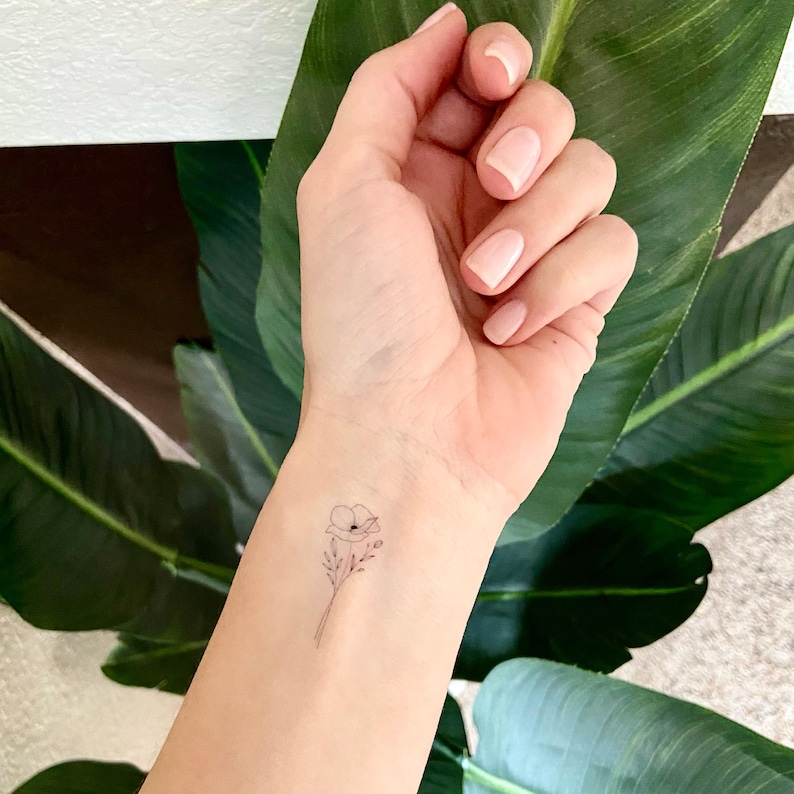 May include: A small, fine-line temporary tattoo of a single poppy flower with delicate stems and leaves applied to the inner wrist. The design features thin, black ink outlines on light skin, showcasing a minimalist botanical illustration style.