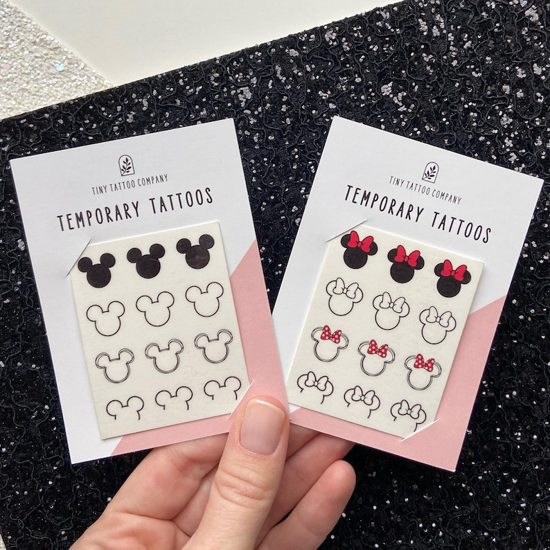 May include: Two white rectangular cards labelled Tiny Tattoo Company Temporary Tattoos. The left card features twelve black mouse-ear silhouettes in solid, outline, and double-line styles. The right card displays twelve mouse-ear designs with red bows.