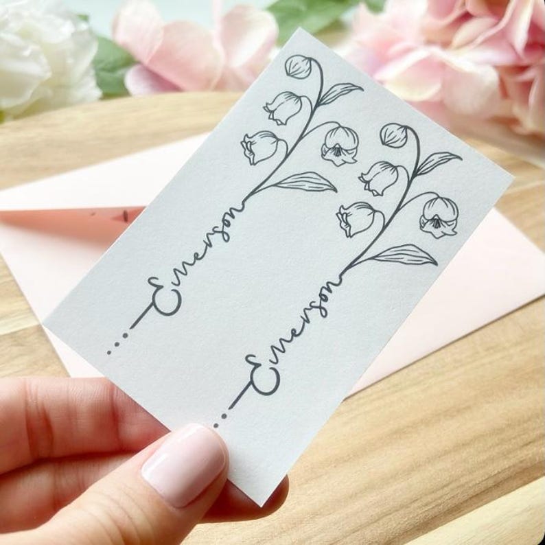 May include: A rectangular white card featuring two identical black line art illustrations of lily of the valley flowers. Each floral stem is integrated with the name Emerson written in a cursive script, ending with three small dots at the base.