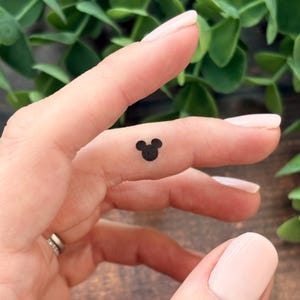 May include: A small, black Mickey Mouse head tattoo on a finger. The tattoo is a simple, minimalist design, with the iconic three-circle silhouette. The skin tone is fair, and the background is blurred green foliage.