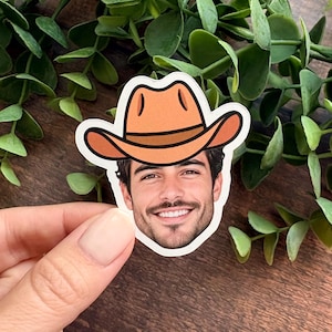 Puede incluir: Una pegatina de vinilo troquelada que presenta un retrato fotogr&aacute;fico realista de una persona sonriente con cabello oscuro y barba, que lleva un sombrero de vaquero ilustrado de color naranja-marr&oacute;n con una banda marr&oacute;n oscuro.
