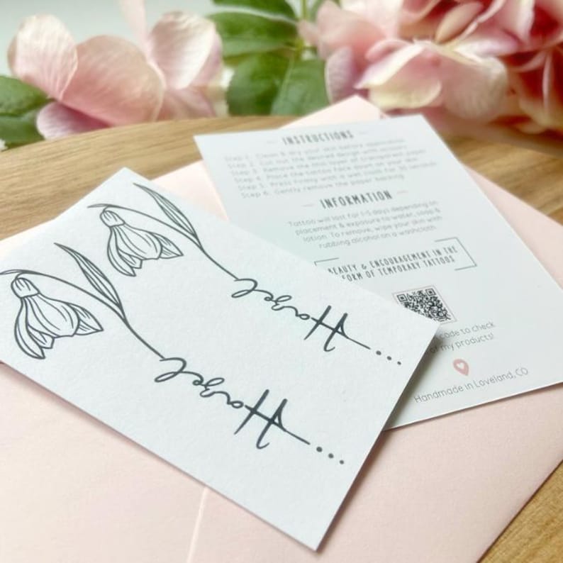 May include: A white rectangular sheet featuring two black line art illustrations of snowdrop flowers with the name Hazel written in cursive script. An accompanying instruction card provides application steps, product information, and a QR code.