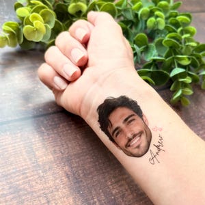 Puede incluir: Un tatuaje temporal aplicado sobre la piel que presenta un retrato realista a color de una persona sonriente con cabello oscuro y barba, acompa&ntilde;ado de dos peque&ntilde;os contornos de corazones rosas y el nombre Andrew en letra cursiva negra.