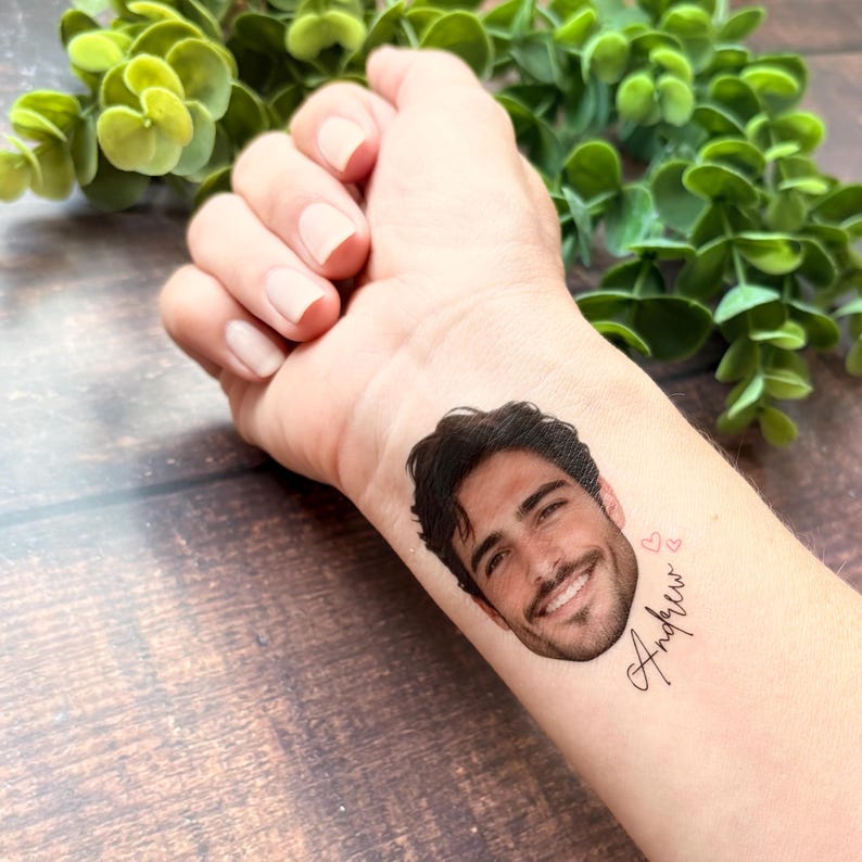 Puede incluir: Un tatuaje temporal aplicado sobre la piel que presenta un retrato realista a color de una persona sonriente con cabello oscuro y barba, acompa&ntilde;ado de dos peque&ntilde;os contornos de corazones rosas y el nombre Andrew en letra cursiva negra.