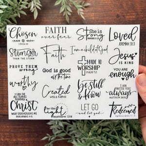 May include: A white card featuring various inspirational quotes in black script. Phrases include "Chosen," "Faith over fear," and "Strong." The card is held by a hand, with greenery in the background.