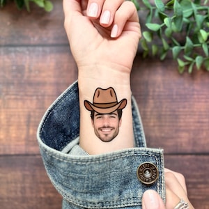 Puede incluir: Un tatuaje temporal aplicado sobre la piel que muestra el retrato realista de una persona sonriente con barba y un sombrero de vaquero marr&oacute;n. El tatuaje est&aacute; situado en la parte interna de la mu&ntilde;eca, visible sobre una manga de chaqueta vaquera.
