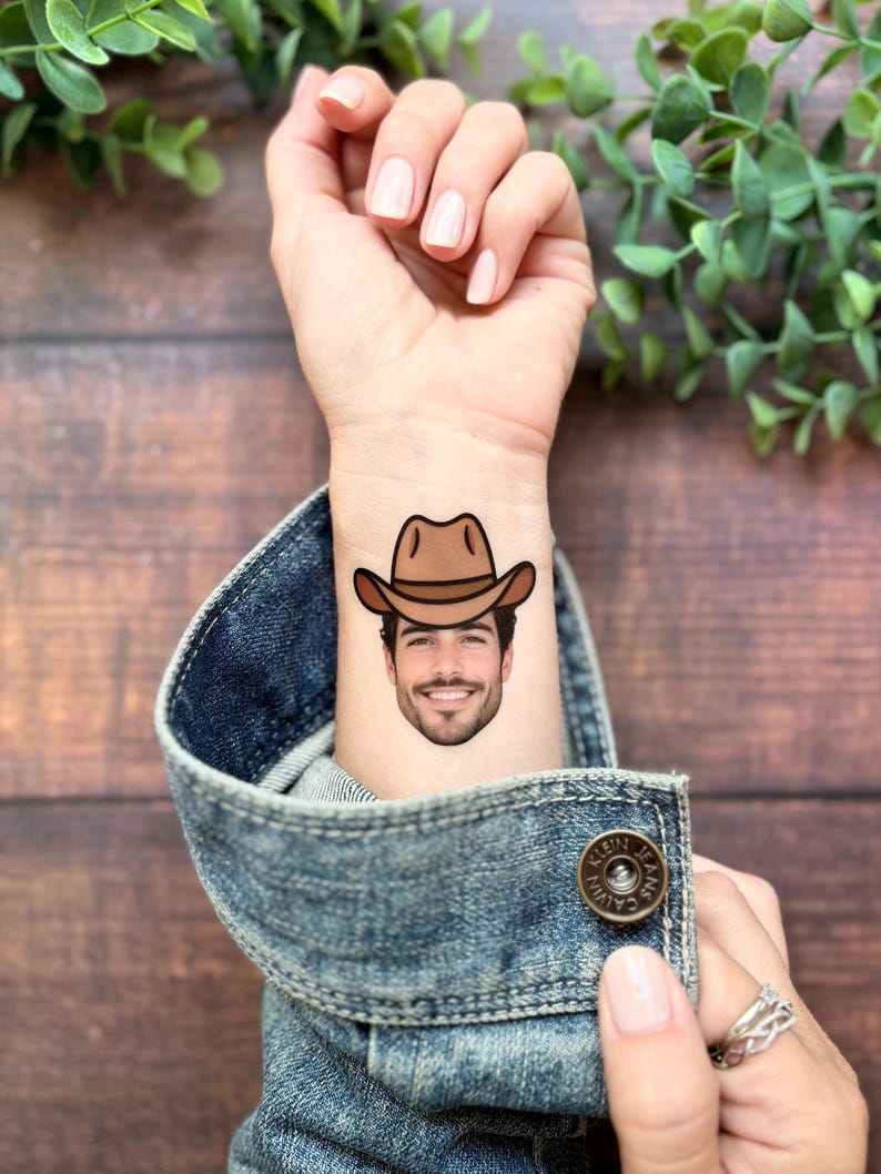 Puede incluir: Un tatuaje temporal aplicado sobre la piel que muestra el retrato realista de una persona sonriente con barba y un sombrero de vaquero marr&oacute;n. El tatuaje est&aacute; situado en la parte interna de la mu&ntilde;eca, visible sobre una manga de chaqueta vaquera.