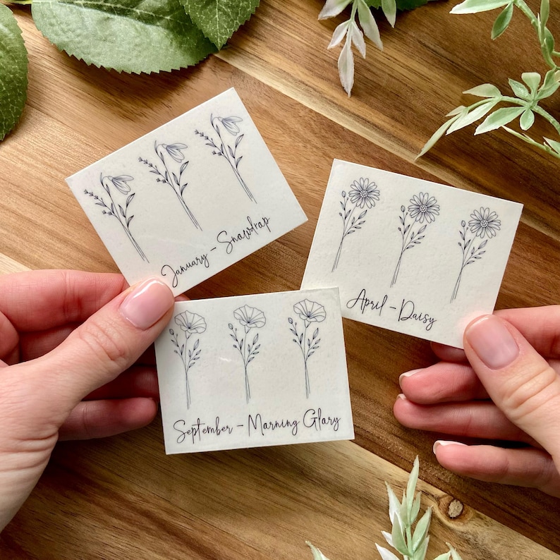 May include: Three rectangular white stickers featuring black line art illustrations of birth flowers. One sticker shows snowdrops labeled January, one shows daisies labeled April, and one shows morning glories labeled September in cursive script.