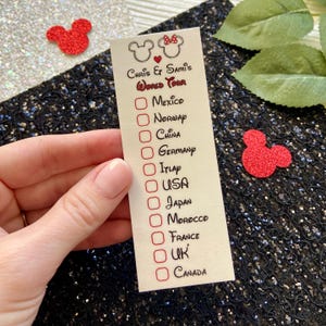 May include: A rectangular off-white checklist titled Chris & Sami's World Tour with two mouse-ear outlines and a red heart. The list includes Mexico, Norway, China, Germany, Italy, USA, Japan, Morocco, France, UK, and Canada with red checkboxes.