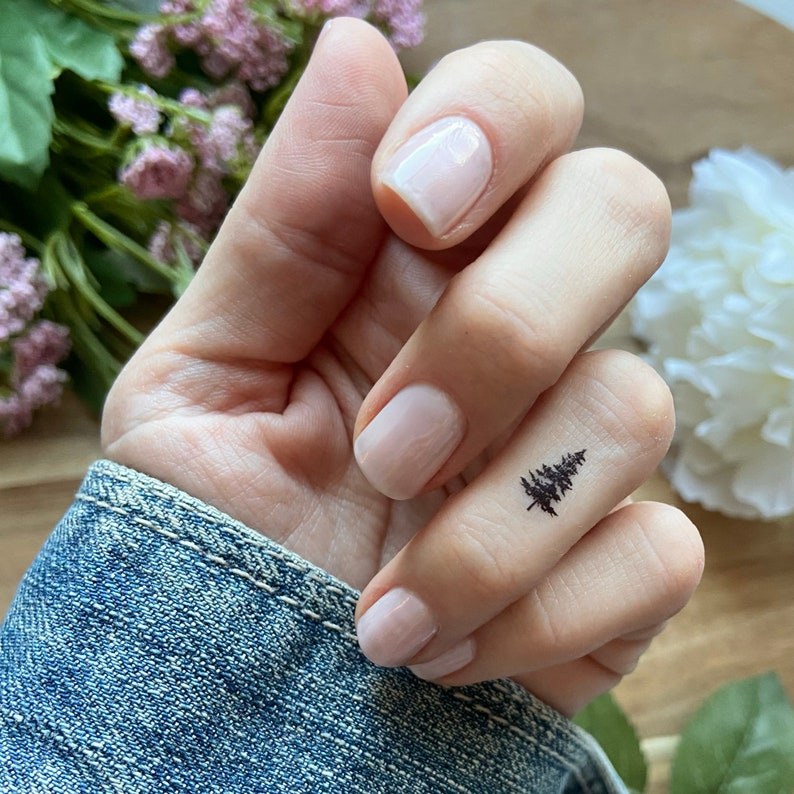May include: A black temporary tattoo of a pine tree on a finger. The hand has manicured nails and is wearing a denim jacket. Flowers are visible in the background.