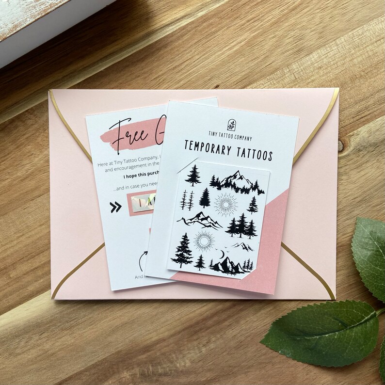 May include: A pink envelope with gold trim holds two cards. One card reads "Free Gift" and the other says "TEMPORARY TATTOOS" with a mountain and tree design. The temporary tattoos are black and white.
