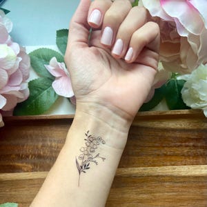 Puede incluir: Un tatuaje temporal que presenta una delicada ilustraci&oacute;n de l&iacute;neas negras de una rama de lirio de los valles con peque&ntilde;as flores en forma de campana y hojas delgadas, aplicado en la mu&ntilde;eca con un dise&ntilde;o bot&aacute;nico limpio y preciso.