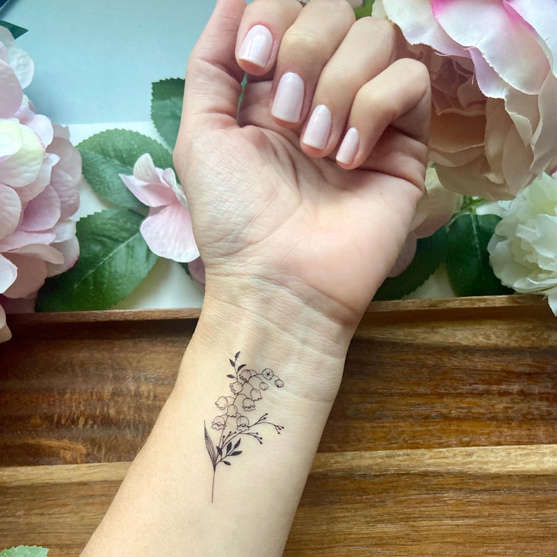 Puede incluir: Un tatuaje temporal que presenta una delicada ilustraci&oacute;n de l&iacute;neas negras de una rama de lirio de los valles con peque&ntilde;as flores en forma de campana y hojas delgadas, aplicado en la mu&ntilde;eca con un dise&ntilde;o bot&aacute;nico limpio y preciso.
