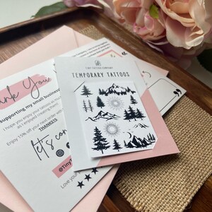 May include: A package of temporary tattoos featuring mountain, sun, and tree designs. The package is on a pink card with the text "TEMPORARY TATTOOS". A card with the text "ink You" and "supporting my small business" is also visible.