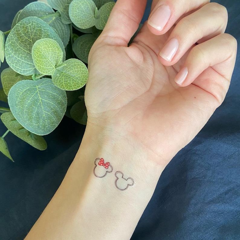 May include: A temporary tattoo on a wrist featuring two black outline silhouettes of mouse ears. One silhouette includes a small red bow with white polka dots positioned between the ears. The design is simple, clean, and minimalist in its execution.