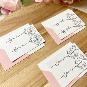 May include: Three white rectangular cards with black ink illustrations of flowers and the names Brecken, Hazel, and Emerson printed twice on each card. Each card is placed on a light pink envelope, featuring delicate line art floral designs.