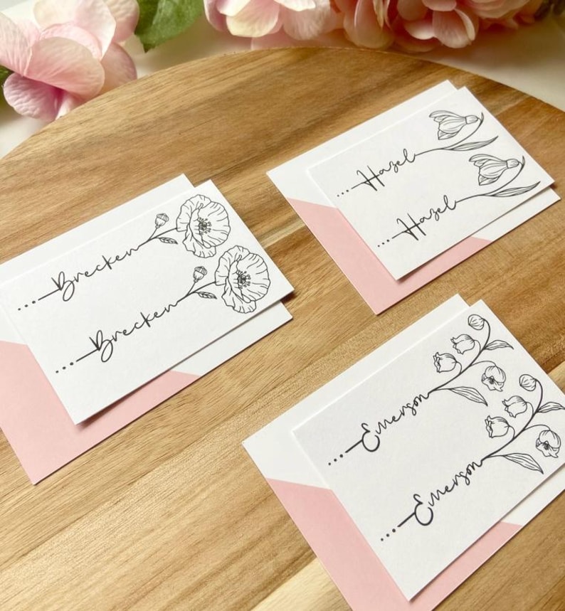 May include: Three white rectangular cards with black ink illustrations of flowers and the names Brecken, Hazel, and Emerson printed twice on each card. Each card is placed on a light pink envelope, featuring delicate line art floral designs.