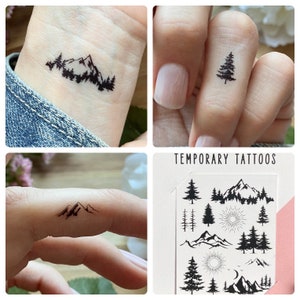 May include: A collection of temporary tattoos featuring mountain and tree designs. The tattoos are black and applied to skin, showcasing the designs on a wrist and fingers. The sheet of tattoos includes various mountain and forest scenes.