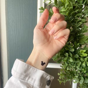 May include: A small, black, circular silhouette of a mouse head icon applied as a temporary tattoo on the inner wrist. The design features one large central circle with two smaller circles positioned symmetrically on top, resembling ears.