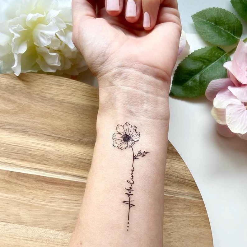 May include: A temporary tattoo on a forearm featuring a fine-line black ink drawing of a single flower with a stem that transitions into the cursive script name Addeline, ending with three small dots aligned vertically below the text.