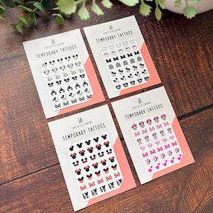 May include: Four white rectangular cards labeled Tiny Tattoo Company Temporary Tattoos, each featuring a grid of small black, pink, and red adhesive designs including mouse ears, bows, castles, diamonds, hearts, and the word love in various styles.