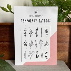 May include: A white card with the text "TINY TATTOO COMPANY TEMPORARY TATTOOS" and a sheet of temporary tattoos featuring black line drawings of various leaves and sprigs. The card has a pink accent on the corner.