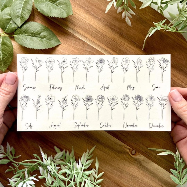 May include: A rectangular white card featuring black line art illustrations of twelve distinct flowers, each labeled with a month from January through December in a cursive script font, arranged in two horizontal rows of six floral designs.